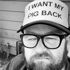 A bearded man with dark rimmed glasses scowls while wearing a hat that reads 'I want my pig back'